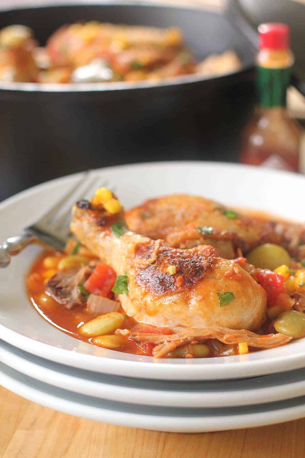 A plate of Brunswick Stew, featuring stewed chicken with corn, lima beans, tomatoes, and herbs, sits on stacked white plates, with a fork and hot sauce bottle nearby.