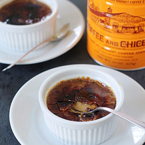 Two white ramekins of crème brûlée, each on a white plate with a spoon. One crème brûlée has a spoon breaking its caramelized sugar top. A Café Du Monde coffee can is in the background.