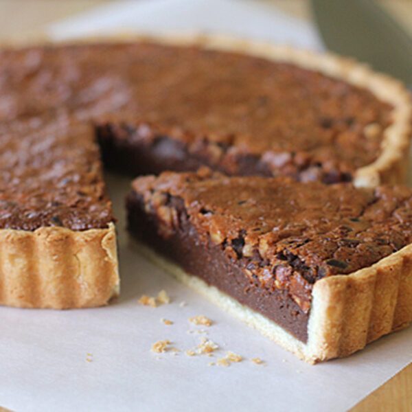 A chocolate tart with a golden crust sits on parchment paper, with one slice cut and slightly pulled out, showing its rich, fudgy filling. A pie server and knife are visible in the background.