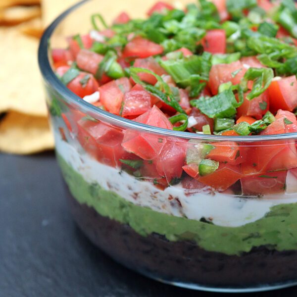 A glass bowl filled with layered dip, including black beans, guacamole, sour cream, diced tomatoes, and chopped green onions. Tortilla chips are visible in the background on a dark surface.