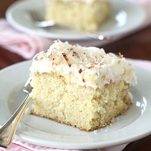 A slice of coconut cake with white frosting and shredded coconut on top sits on a white plate with a fork, atop a pink napkin; another slice is blurred in the background.