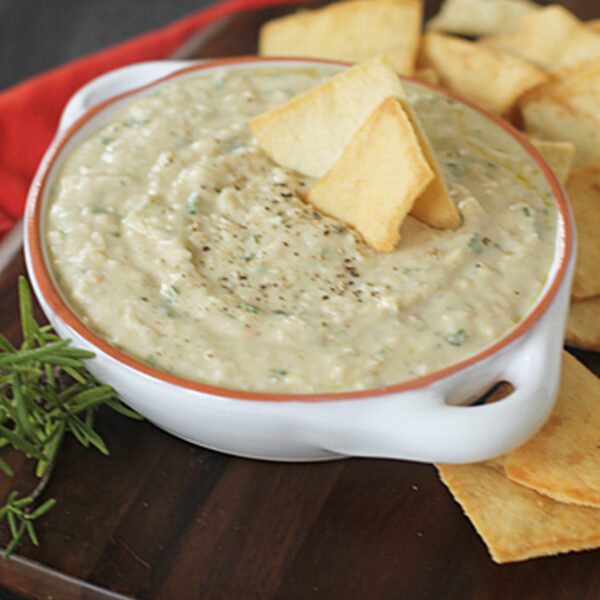 A bowl of creamy dip topped with cracked black pepper and served with pita chips on a wooden board, garnished with a sprig of fresh herbs.