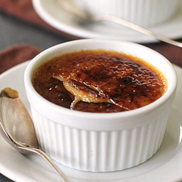 A white ramekin filled with crème brûlée sits on a white plate with a silver spoon. The caramelized sugar top is cracked, revealing the creamy custard beneath. Another ramekin of crème brûlée is visible in the background.