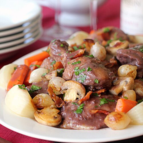 A plate of coq au vin with chicken, mushrooms, pearl onions, carrots, and bacon, garnished with parsley. Wine glasses, a wine bottle, plates, and a gravy boat are in the blurred background.