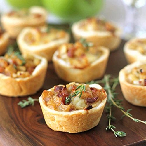 Mini savory tarts filled with caramelized onions and herbs are arranged on a wooden board, with fresh thyme sprigs and green apples in the background.