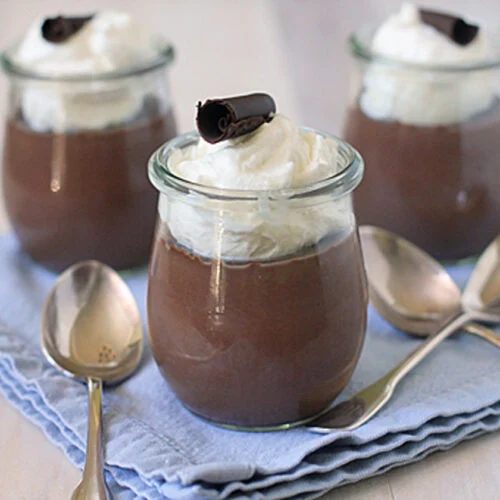 Three glass jars filled with chocolate mousse, topped with whipped cream and a chocolate curl, are placed on a folded blue cloth, surrounded by three metal spoons.