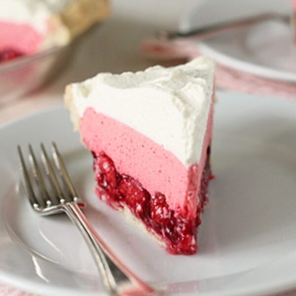 A slice of pie with a layer of red berry filling, a thick layer of pink mousse, and a topping of whipped cream sits on a white plate next to a fork.