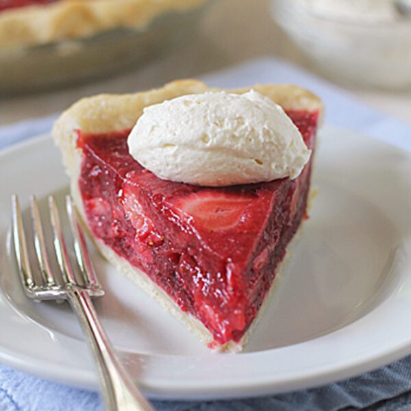 A slice of strawberry pie topped with a dollop of whipped cream sits on a white plate with a fork, with the rest of the pie and a bowl of whipped cream in the background.