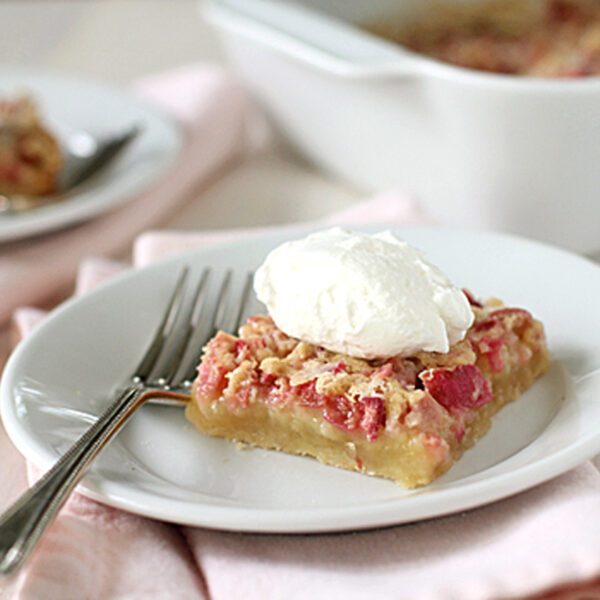 A slice of dessert bar topped with whipped cream sits on a white plate with a fork, resting on a pale pink napkin. Another plate and a baking dish can be seen in the background.