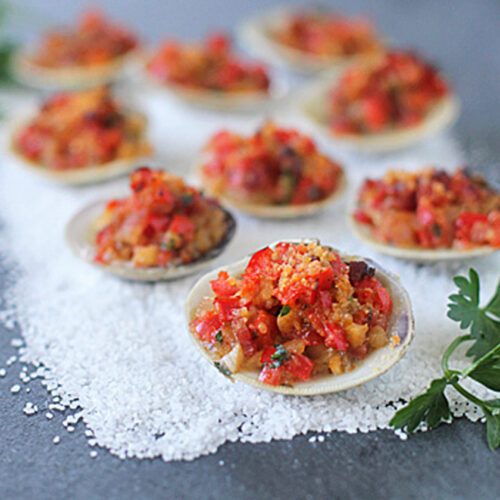 A close-up of stuffed clams topped with a colorful mixture of breadcrumbs, herbs, and diced vegetables, arranged on a bed of coarse salt with fresh parsley garnishes.