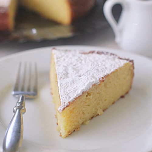 A slice of yellow cake dusted with powdered sugar sits on a white plate with a fork. A small pitcher and the rest of the cake on a platter are in the background.