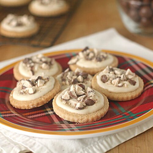 A red plaid plate with six frosted cookies topped with chopped chocolate pieces sits on a white napkin. More cookies and a glass bowl of chocolates are in the blurred background.