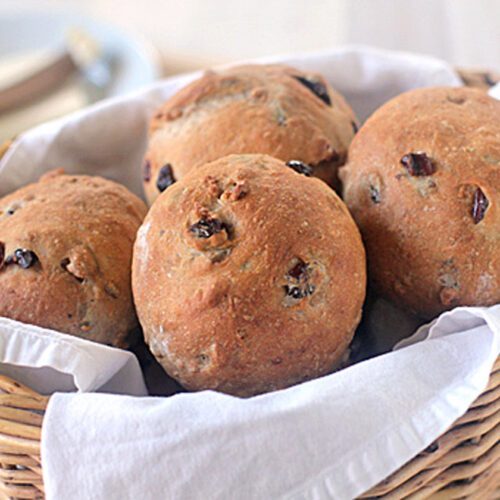 A wicker basket lined with a white cloth holds four round, golden-brown raisin bread rolls with visible raisins baked into the surface. The background is softly blurred with tableware visible.