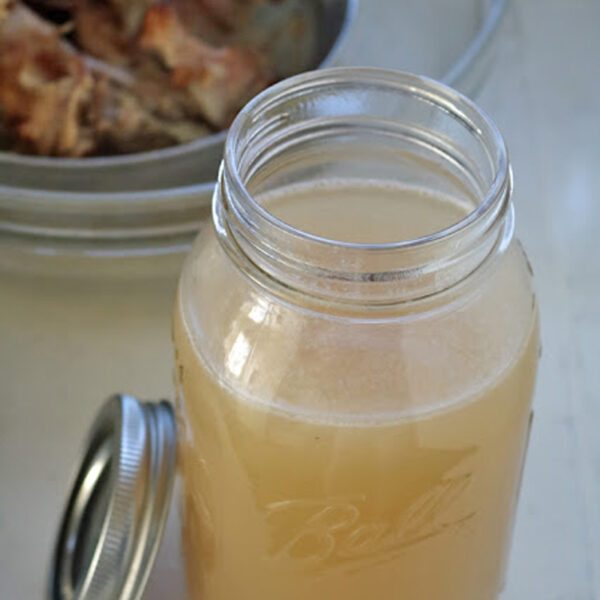 A glass mason jar filled with light brown homemade broth sits on a countertop with its lid off. In the background, a metal bowl holds cooked meat and bones.