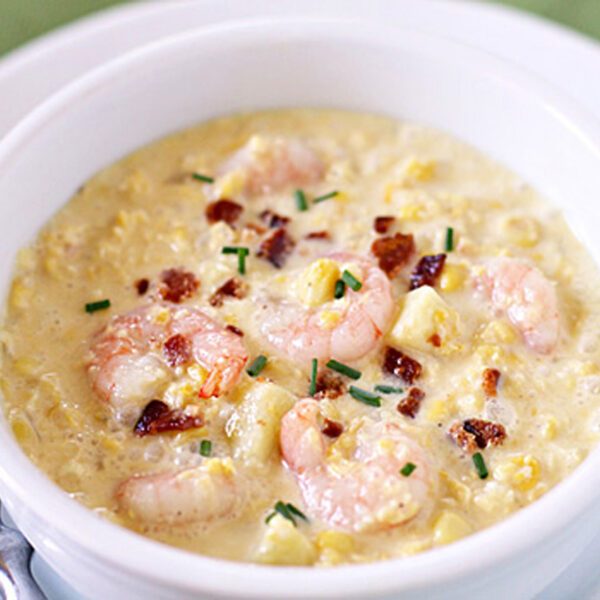 A white bowl filled with creamy shrimp chowder, garnished with crumbled bacon and chopped chives, sits next to a silver spoon on a white plate.