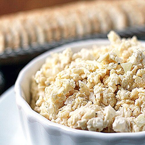 A white bowl filled with chunky homemade pimento cheese spread sits on a plate, with a stack of crackers blurred in the background.
