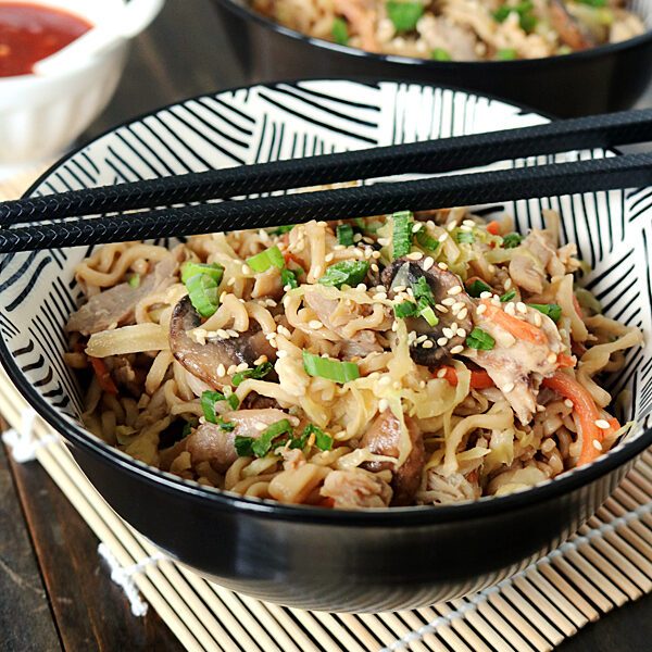 A bowl of stir-fried noodles with vegetables, mushrooms, and sesame seeds, garnished with chopped green onions. Black chopsticks rest on the bowl, and a small dish of red sauce is in the background.