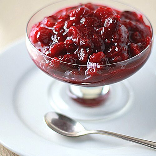 A glass dish filled with chunky cranberry sauce sits on a white plate with a silver spoon beside it.