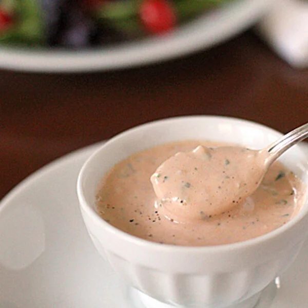 A small white bowl filled with creamy pink salad dressing, garnished with herbs, sits on a white plate. A spoon rests in the bowl, with some dressing on it. A blurry salad appears in the background.