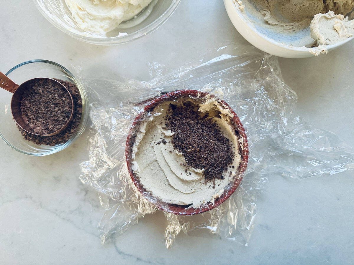 A bowl lined with plastic wrap contains a creamy mixture topped with grated chocolate. Nearby are a small glass bowl with more chocolate shavings and another bowl with leftover cream on a light countertop.