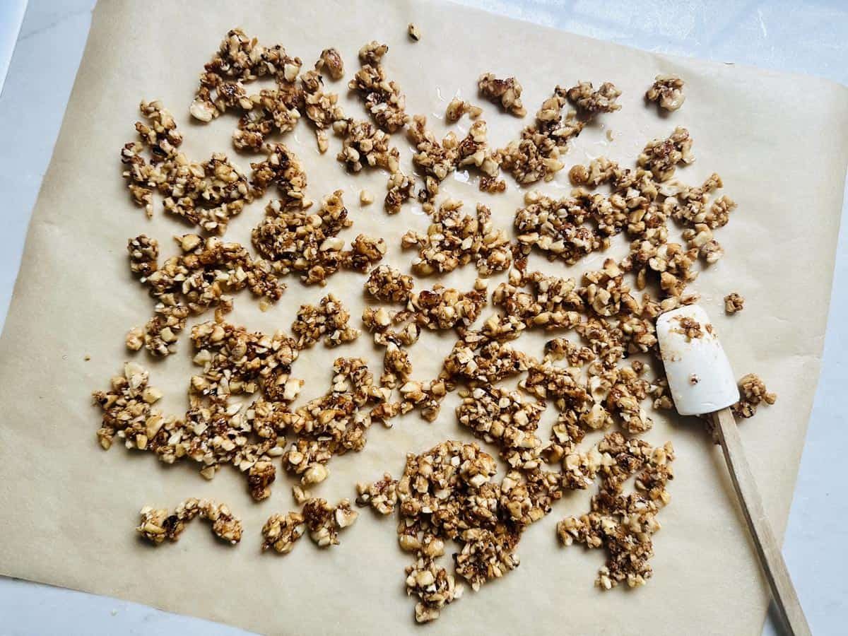 Clusters of nut brittle are scattered on a sheet of parchment paper, with a white spatula resting next to them. The brittle appears golden brown and chunky, suggesting a homemade mixture.