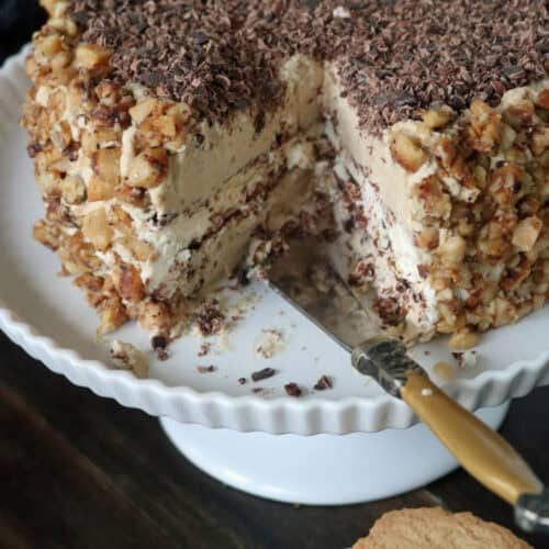 A layered cake with creamy frosting, chopped nuts on the sides, and chocolate shavings on top sits on a white cake stand. A slice has been cut, revealing the inner layers, with a knife beside the cake.