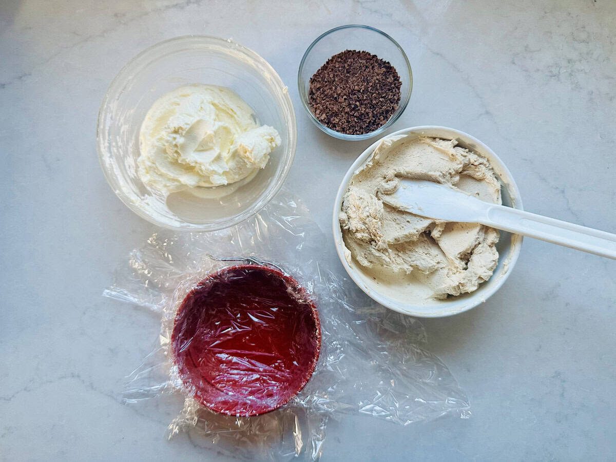 A countertop displays a bowl of whipped cream, a bowl of coffee ice cream with a spatula, a small bowl of ground coffee, and a red bowl lined with plastic wrap.