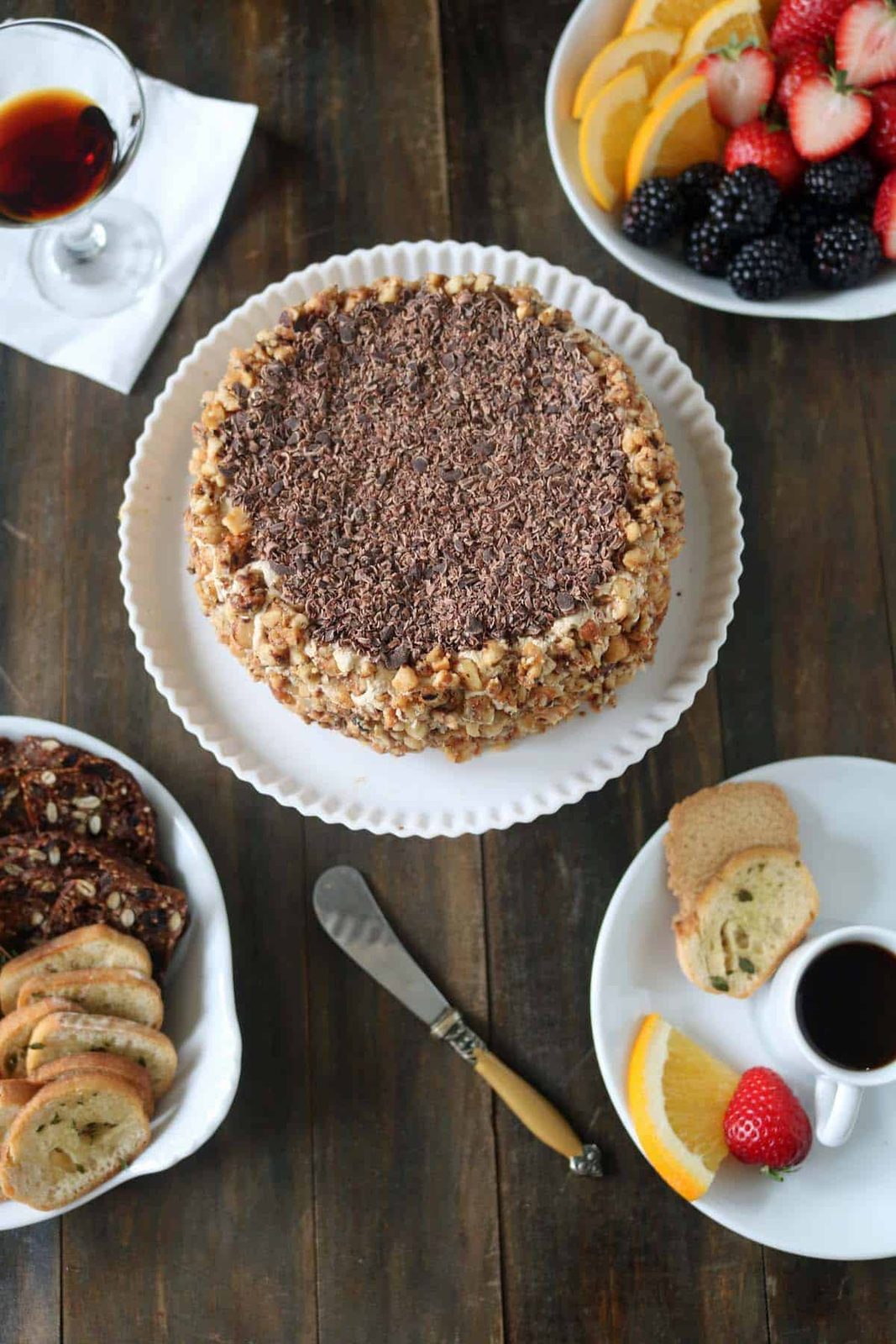 A round cake topped with chocolate shavings and chopped nuts sits on a white plate, surrounded by a fruit platter, biscotti, a cup of coffee, and a glass of liqueur on a wooden table.