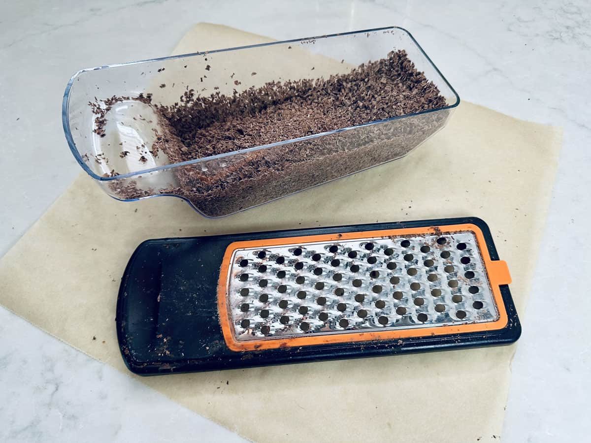 A box grater with grated chocolate inside and some chocolate shavings scattered around, placed on a sheet of parchment paper on a light countertop.
