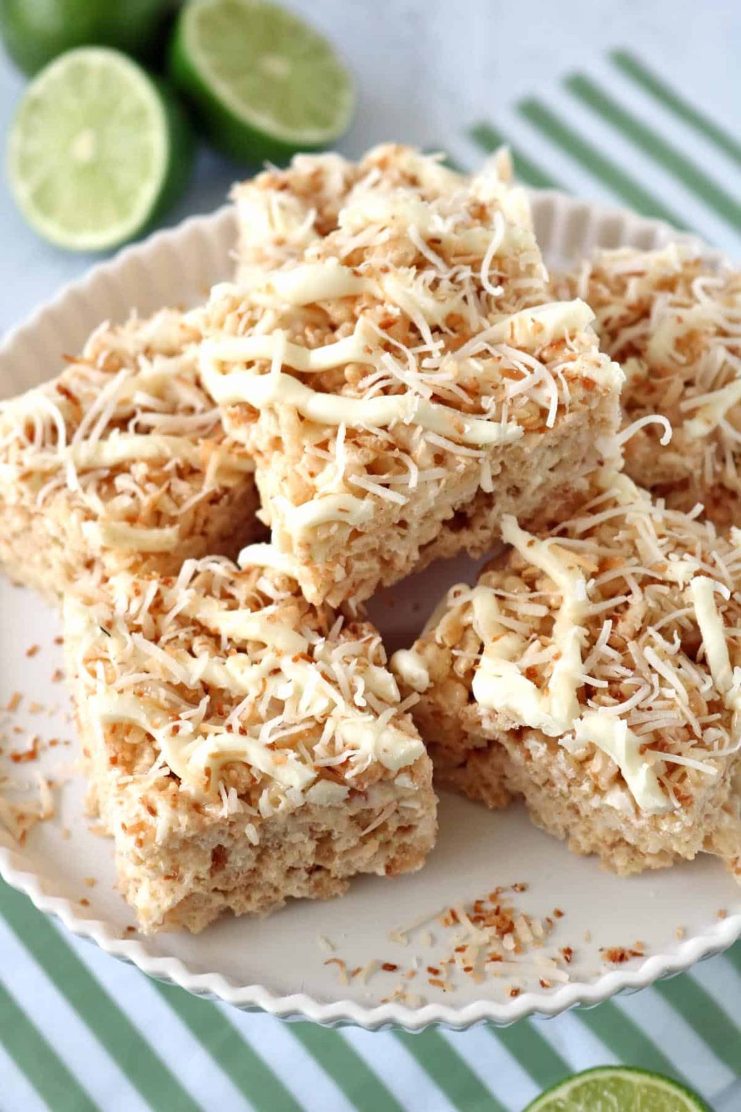 A plate of coconut Rice Krispies treats topped with toasted coconut and white chocolate drizzle, with sliced limes in the background on a green-striped cloth.