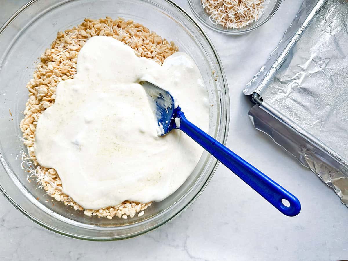 A bowl of rice cereal topped with melted marshmallow mixture is being mixed with a blue spatula. Next to it are a small bowl of toasted coconut and a foil-lined baking pan.