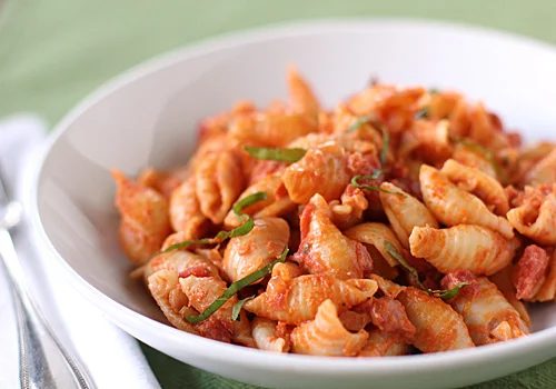 A white bowl filled with shell-shaped pasta covered in tomato sauce, garnished with thin strips of fresh basil, sits on a green surface next to a fork and spoon.