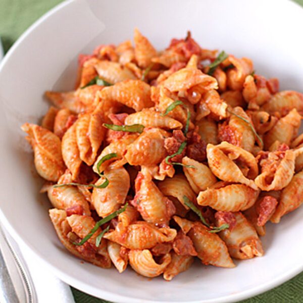 A white bowl filled with shell pasta topped with tomato sauce and garnished with fresh basil, placed on a green tablecloth next to a fork and napkin.
