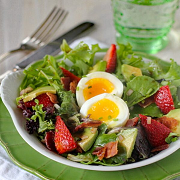 A fresh salad with mixed greens, avocado slices, strawberries, crispy bacon pieces, and a soft-boiled egg, served on a white plate with a green rim. A fork, knife, and clear green glass are nearby.
