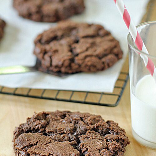 A close-up of a large, chocolate cookie on a wooden surface, with a glass of milk and a red-striped straw beside it. More cookies are cooling on a rack lined with parchment paper in the background.