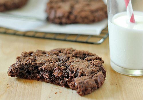 A chocolate cookie with a bite taken out is on a wooden surface next to a glass of milk with a red-striped straw. More cookies are visible on a cooling rack in the background.