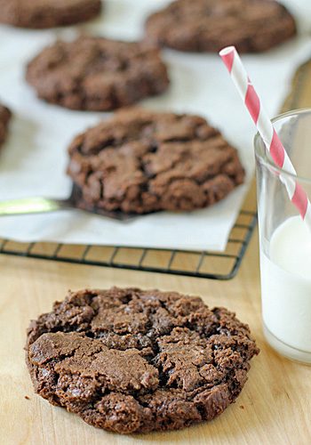 A thick chocolate cookie rests on a wooden surface next to a glass of milk with a red and white striped straw. More cookies cool on a wire rack lined with parchment paper in the background.