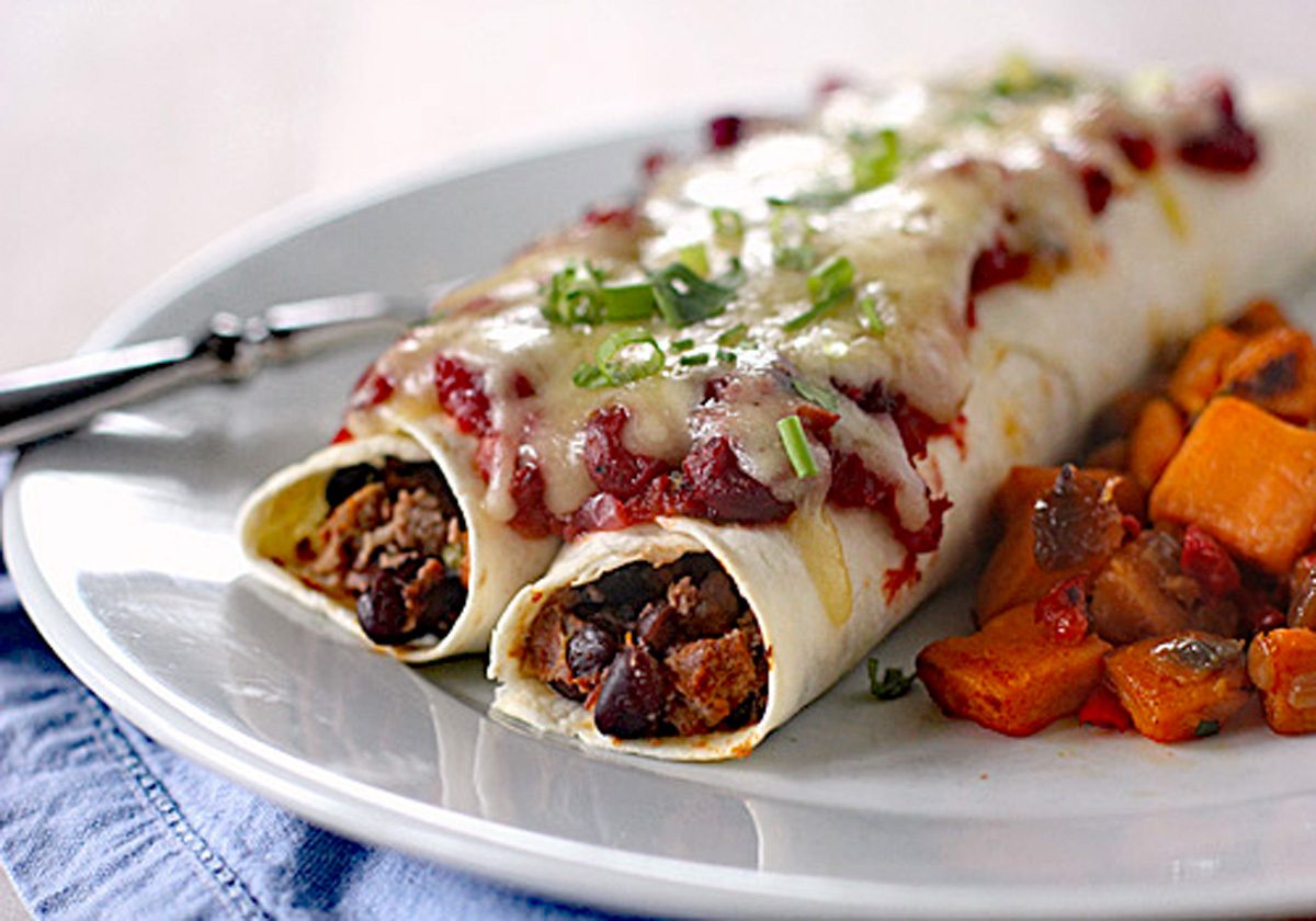 Two enchiladas filled with beans and meat, topped with melted cheese, tomato sauce, and chopped green onions, served on a white plate with a side of roasted vegetables.