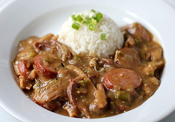 A plate of gumbo with sliced sausage, shredded meat, and vegetables in a brown sauce, served with a mound of white rice topped with chopped green onions.