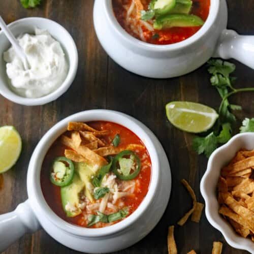 Two bowls of tortilla soup topped with avocado slices, jalapeño, cheese, and tortilla strips on a wooden table, surrounded by lime wedges, cilantro, sour cream, and extra tortilla strips.