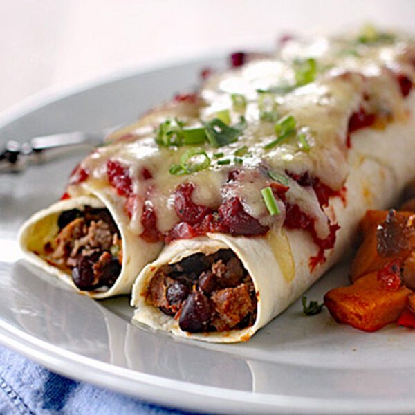 Two enchiladas filled with beans and meat, topped with melted cheese, tomato sauce, and chopped green onions, served on a white plate with a side of roasted vegetables.