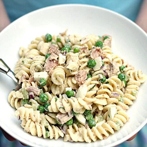 A person holds a white bowl filled with rotini pasta salad, featuring green peas, chunks of tuna, and diced vegetables, with a fork resting inside the bowl.