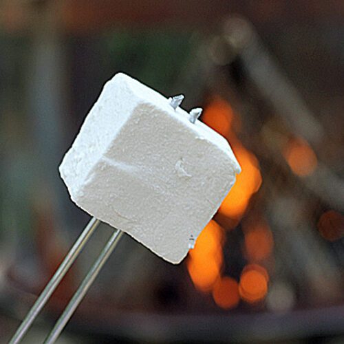 A close-up of a marshmallow on a metal skewer held in front of a campfire with orange flames and logs in the background.