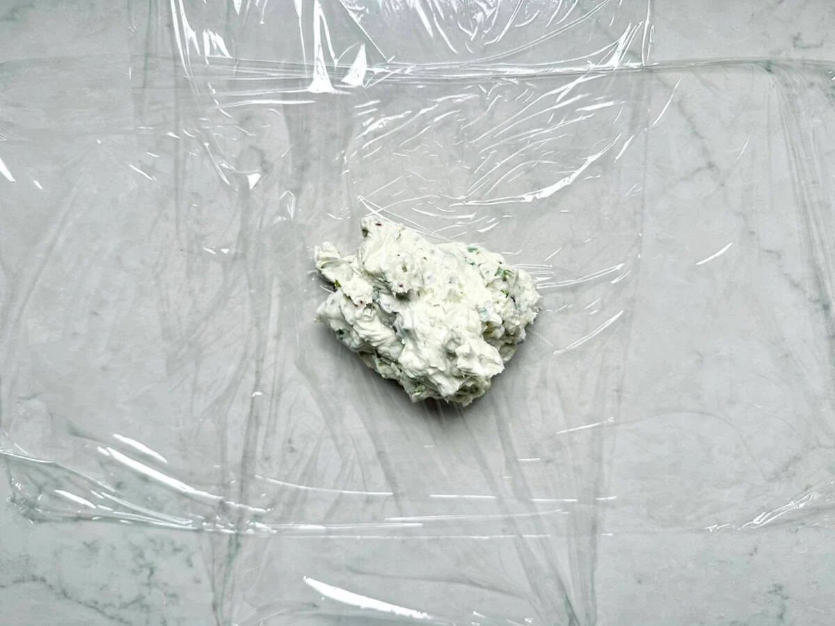 A dollop of herbed cream cheese, inspired by Vivian Howard’s Party Magnet, sits on plastic wrap atop a marble surface, ready to be wrapped.