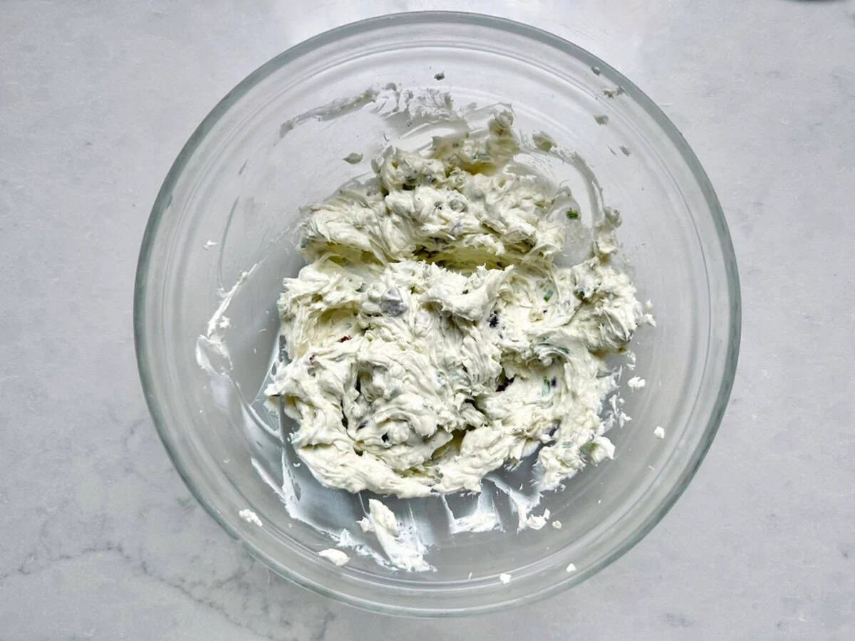 Glass bowl with herbed cream cheese mixture on a light marble surface; flecks of green herbs and seasoning are visible throughout the creamy, spreadable mixture—just like Vivian Howard’s Party Magnet, it’s irresistibly inviting and perfect for any gathering.
