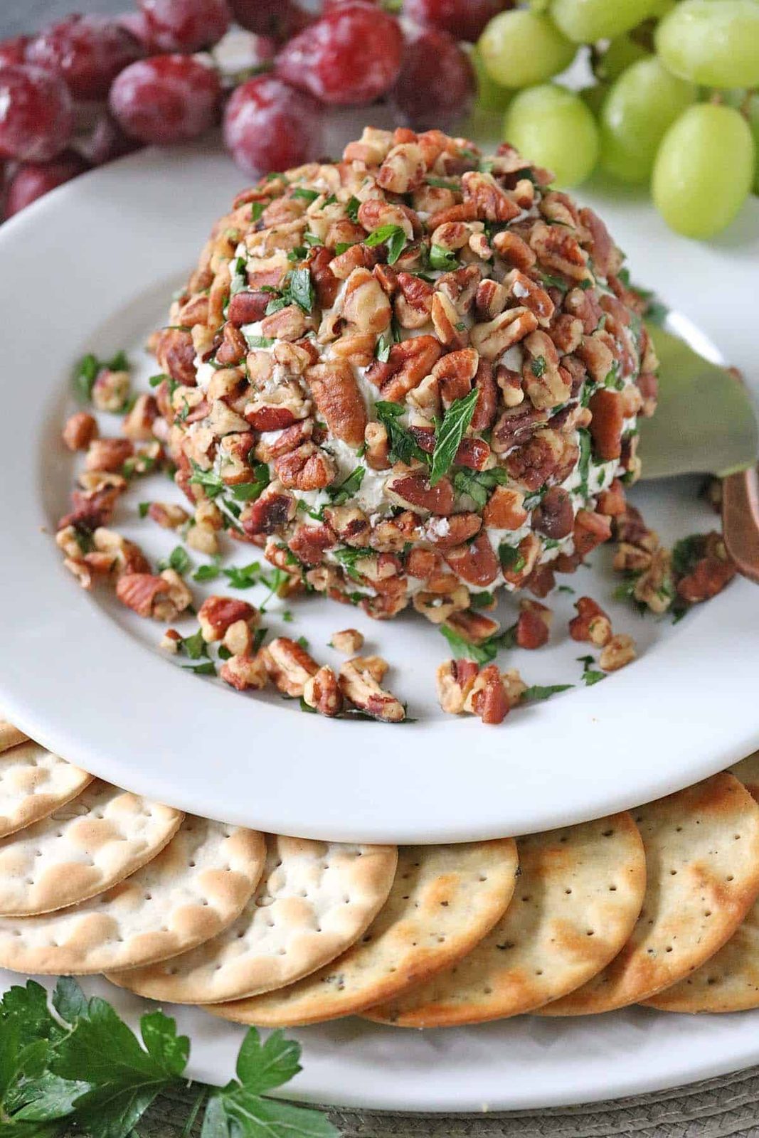 A cheese ball—Vivian Howard’s Party Magnet—coated with chopped pecans and herbs sits on a white plate, surrounded by round crackers and red and green grapes.