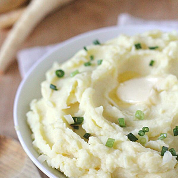 A bowl of creamy mashed potatoes topped with chopped green onions and a melting pat of butter, placed on a wooden surface with a wooden spoon nearby.
