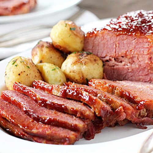 Sliced glazed corned beef with a side of seasoned roasted potatoes on a white plate, served with silverware, and a blurred background plate visible.