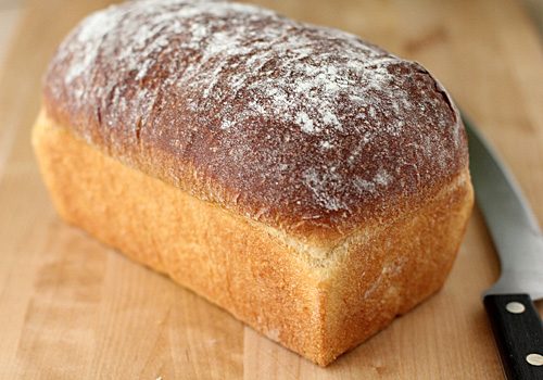 A freshly baked loaf of bread with a golden-brown crust, lightly dusted with flour, sits on a wooden surface next to a bread knife.