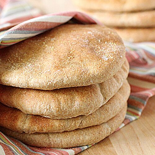 A stack of round, golden-brown pita breads rests partially wrapped in a colorful striped cloth on a wooden surface. More pita breads are visible in the background.